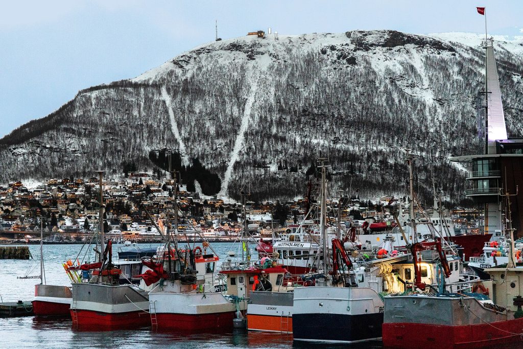 Kalastusveneitä Tromssan satamassa talvella, taustalla luminen Tromsdalen ja Fjellheisenin köysiradan reitti